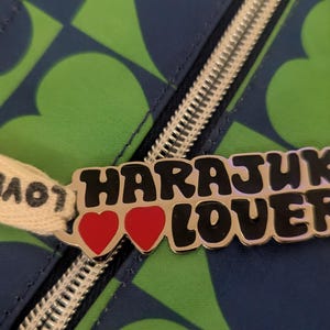 May include: Close-up of a zipper pull with the words "Harajuku Lovers" in black and silver. The pull features two red hearts and is attached to a blue and green heart-patterned fabric.