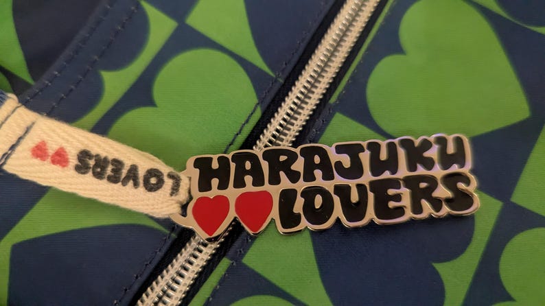 May include: Close-up of a zipper pull with the words "Harajuku Lovers" in black and silver. The pull features two red hearts and is attached to a blue and green heart-patterned fabric.