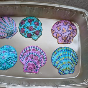 Puede incluir: Seis conchas marinas pintadas a mano en una bandeja plateada. Las conchas están decoradas con varios diseños, incluyendo corazones, rosas y patrones abstractos, en tonos de rosa, azul y morado. La bandeja es rectangular.