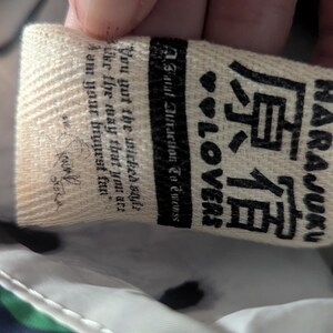 May include: Close-up of a cream-colored tag with the text "HARAJUKU" in English and Japanese characters. The tag also includes the words "LOVERS" and a quote. The background is a patterned fabric.