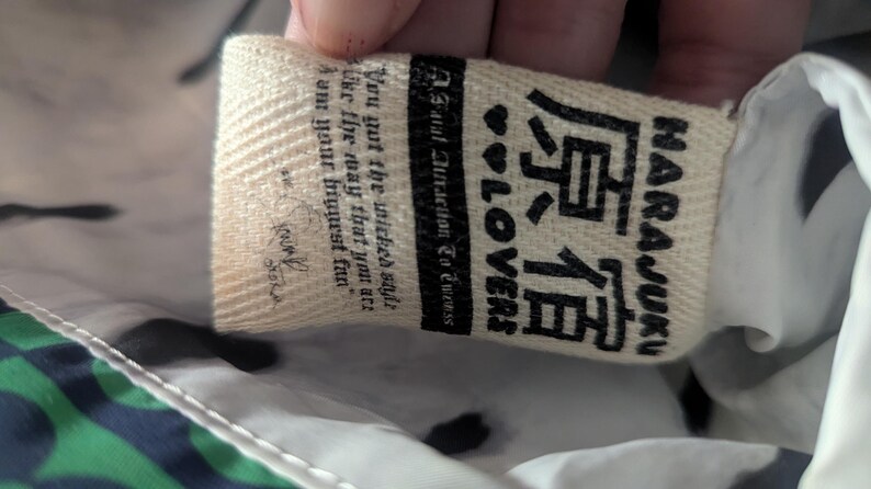 May include: Close-up of a cream-colored tag with the text "HARAJUKU" in English and Japanese characters. The tag also includes the words "LOVERS" and a quote. The background is a patterned fabric.