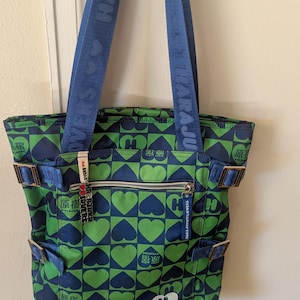 May include: A green and blue tote bag with a heart pattern. The bag has blue straps with white text and multiple pockets. The bag is hanging on a door.