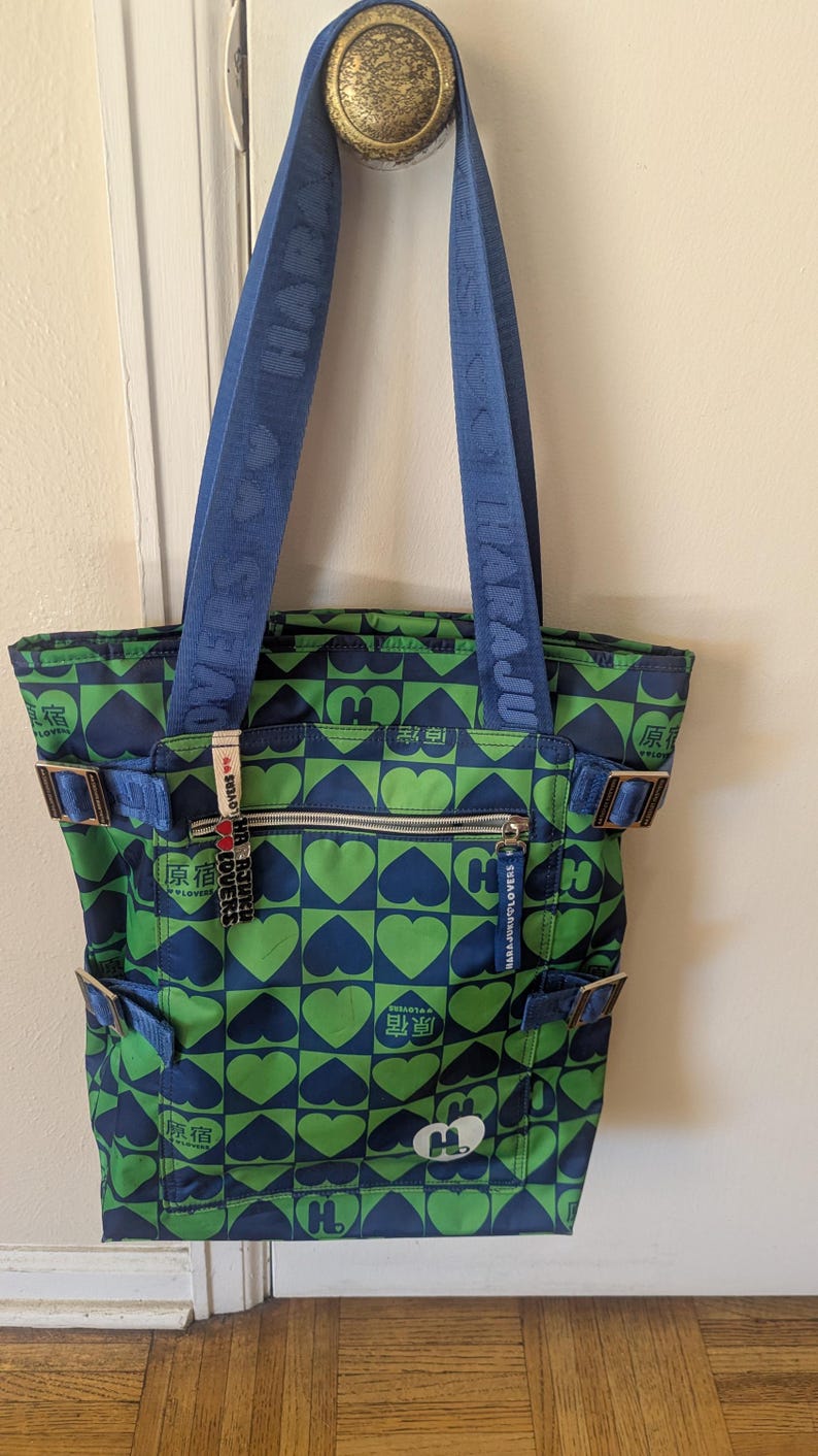 May include: A green and blue tote bag with a heart pattern. The bag has blue straps with white text and multiple pockets. The bag is hanging on a door.