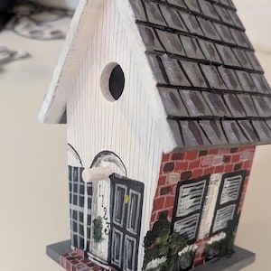 Peut inclure: Nichoir en bois peint à la main en forme de maison. Il présente une façade blanche avec un trou d'entrée rond, un toit en bardeaux gris et une base en forme de brique. La porte d'entrée porte les numéros 1, 2 et 3.