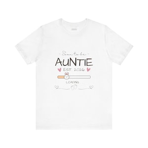 May include: White t-shirt with the text "Soon to be AUNTIE EST. 2026" in a script font. The design includes pink hearts, a loading bar with a bunny, and baby footprints. A cute and fun design.