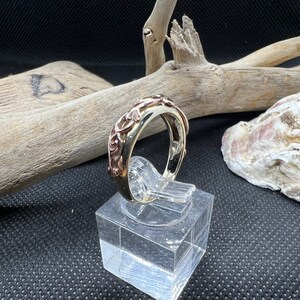 May include: A silver ring with a rose gold band and intricate floral design. The ring is displayed on a clear acrylic stand.