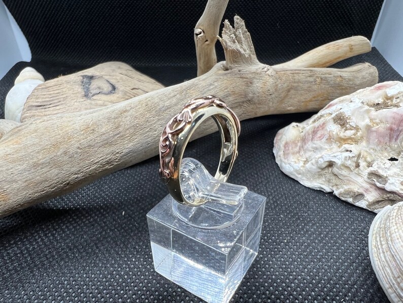 May include: A silver ring with a rose gold band and intricate floral design. The ring is displayed on a clear acrylic stand.