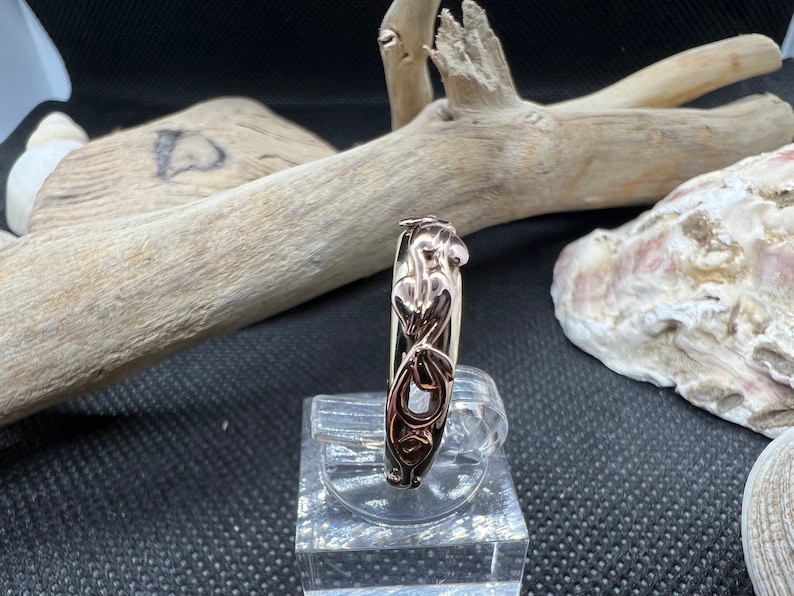 May include: A gold ring featuring an intricate design of interlaced hearts and leaves. The ring is crafted from two distinct shades of gold, resulting in a sophisticated and stylish piece.