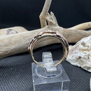 May include: A gold and rose gold band ring with a textured design. The ring is on a display stand.