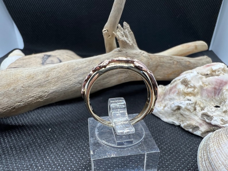 May include: A gold and rose gold band ring with a textured design. The ring is on a display stand.