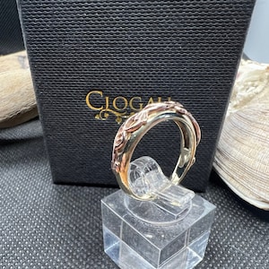 May include: A silver and rose gold ring with a Celtic knot design. The ring is on a white display stand. The ring is in a black box with the word "Ciogail" on it.