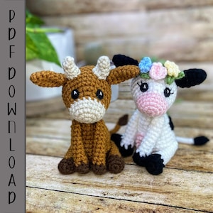 May include: Two crocheted stuffed animals, a brown cow and a white cow with black spots, sitting on a wooden surface. The white cow has a flower crown on its head.