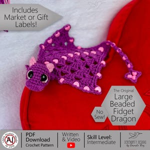 May include: A handmade purple crochet dragon with pink heart accents and large black eyes. The dragon has a long tail and is labeled "The Original Large Beaded Fidget Dragon". The image also includes the text "No Sew!" and "PDF Download Crochet Pattern".