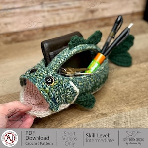 May include: A handmade crochet fish-shaped basket in shades of green, blue, and white, with a beige interior. The basket holds a wallet, keys, and pens. The text "PDF Download Crochet Pattern" is visible.