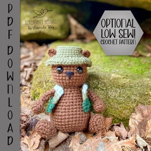 Pu&ograve; includere: Un orso di peluche marrone che indossa un gilet verde e blu e un cappello verde. L'orso &egrave; seduto su un letto di foglie marroni. Il testo "OPTIONAL LOW SEW! CROCHET PATTERN" &egrave; nell'angolo in alto a destra dell'immagine.
