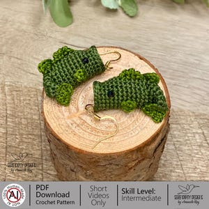 May include: Handmade green crochet dinosaur earrings with gold-colored ear wires. The earrings are displayed on a wooden slice. The earrings have black bead eyes and are made with a detailed crochet stitch. The earrings are a unique accessory.