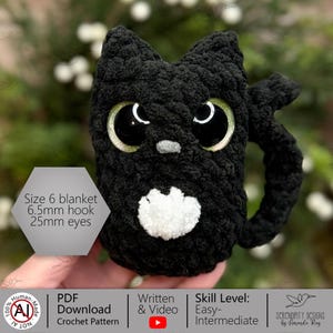 K&ouml;nnte beinhalten: Eine handgefertigte, schwarze H&auml;kelkatzen-Tasse mit gro&szlig;en, glitzernden gr&uuml;nen Augen und einer wei&szlig;en Brust. Die Tasse hat einen Henkel. Das Bild enth&auml;lt den Text "Size 6 blanket, 6.5mm hook, 25mm eyes".