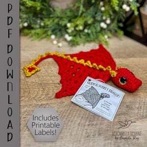 May include: A handmade red crochet dragon with yellow accents and large black eyes. The dragon has a tag that reads "Gliding Pocket Dragon" and "Includes Printable Labels!" on a wooden surface.