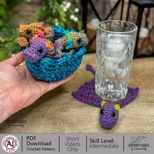 May include: A crocheted basket filled with three colorful dragon figures. The dragons are in shades of orange, purple, and blue. A purple dragon coaster and a glass of water are also visible. The text in the image says "PDF Download Crochet Pattern".