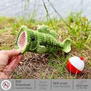 May include: A handmade green crochet fish with an open mouth revealing a pink interior. A black button serves as an eye. A red and white bobber sits nearby on the grass. The image also includes text that says "PDF Download Crochet Pattern".