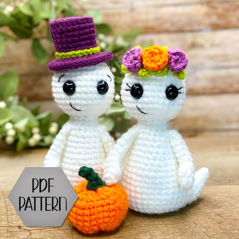 May include: Two crocheted ghost figures, one wearing a purple top hat and the other a floral crown, holding a crocheted orange pumpkin.  PDF PATTERN.