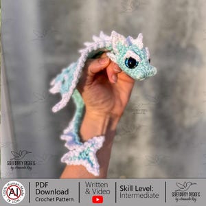 Puede incluir: Un dragón de ganchillo en tonos azules y blancos, sostenido en una mano. El dragón tiene grandes ojos azules y un cuerpo largo y serpentino. El patrón de ganchillo está disponible como descarga en PDF, con instrucciones escritas y en vídeo para un nivel intermedio.