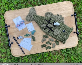 May include: A wooden serving tray displays a collection of handmade crochet items. The centerpiece is a large green fish with a tic-tac-toe game. Other items include fish-shaped earrings, keychains, and small fish. A black pen and tags are also present.
