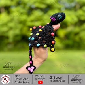 Op de afbeelding: Een handgemaakt zwart gehaakt wezen met kleurrijke kraalaccenten. Het wezen heeft grote blauwe ogen, paarse en roze details en een lange kralenstaart. De afbeelding bevat de tekst "PDF Download Crochet Pattern" en "Skill Level: Intermediate".