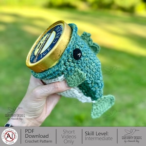 May include: A teal and green crochet fish ice cream cozy, holding a pint of ice cream. The fish has a black button eye and a white belly. The ice cream container has a gold lid. The design is by Serendipity Designs.