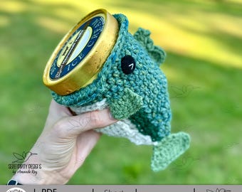 May include: A handmade crochet fish ice cream cozy in shades of teal and green, holding a pint of ice cream. The fish has a black button eye and a white belly. The ice cream container has a gold lid. The design is by Serendipity Designs.