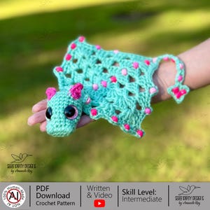 May include: A crocheted dragon toy in shades of teal and pink. The dragon has large, glittery eyes and pink accents on its ears and body. The crochet pattern is available as a PDF download.