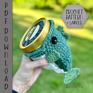 Pode incluir: Padrão de crochê e etiquetas para uma capa de recipiente de sorvete em forma de peixe verde e branco. A capa é feita de crochê e tem um olho preto com um círculo branco ao redor. O texto "CROCHET PATTERN + LABELS" está em um hexágono cinza.