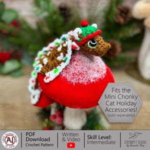 May include: A crocheted cat accessory set, featuring a brown cat head wearing a green and red hat and a gingerbread-style scarf. The cat is perched on a red mushroom-shaped base with a snowy texture. The text says, "Fits the Mini Chonky Cat Holiday Accessories!"