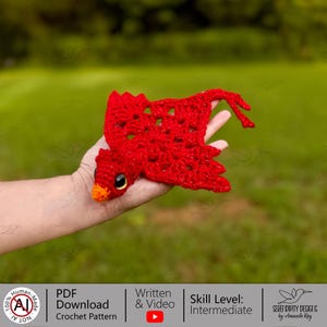 May include: A bright red crocheted bird ornament featuring a textured wing pattern, a small orange beak, and a dark round eye. The bird is crafted with a visible stitch design and has a long, thin tail section extending from the main body.
