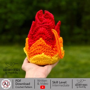 May include: A crocheted flame-shaped object featuring a gradient of colors starting with a yellow base, transitioning to orange in the middle, and topped with bright red textured peaks. The yarn work displays a dense, looped stitch pattern.