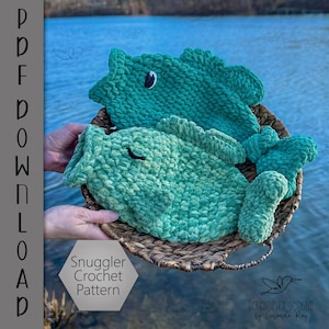 Puede incluir: Dos peces de crochet verde en una cesta de mimbre. El patrón de crochet se llama "Snuggler Crochet Pattern" y está disponible para descargar en formato PDF.