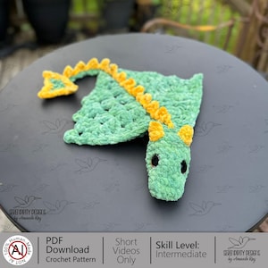 May include: A handmade green crochet dragon with yellow detailing. The dragon features large black eyes and a long tail. The crochet pattern is available as a PDF download. Skill level: Intermediate.