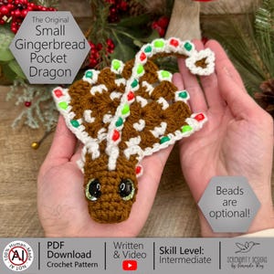 May include: A crocheted gingerbread dragon in brown, white, red, and green. The dragon has large, glittery eyes and is decorated with candy-like embellishments. The image text reads "The Original Small Gingerbread Pocket Dragon."