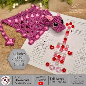 May include: A handmade crochet bookmark in shades of purple, adorned with pink heart-shaped beads. The bookmark features a dragon design with a matching crochet head. A bead planning chart and other craft supplies are also visible.