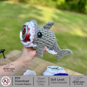 May include: A handmade crochet shark drink cozy in gray, white, and pink, holding a red soda can. The shark has a black button eye and white teeth. The crochet pattern is available as a PDF download.