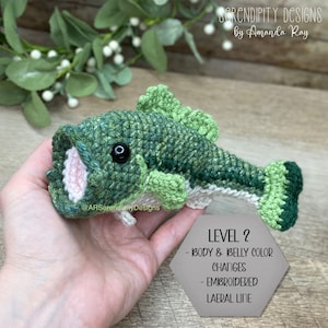 May include: A handmade crochet fish in shades of green, with a white belly and pink mouth. The fish has a black eye and is labeled "LEVEL 2" with details about body and belly color changes and an embroidered lateral line. The text "SERENDIPITY DESIGNS by Amanda Ray" is also visible.