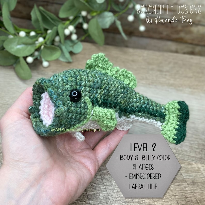 May include: A handmade crochet fish in shades of green, with a white belly and pink mouth. The fish has a black eye and is labeled "LEVEL 2" with details about body and belly color changes and an embroidered lateral line. The text "SERENDIPITY DESIGNS by Amanda Ray" is also visible.