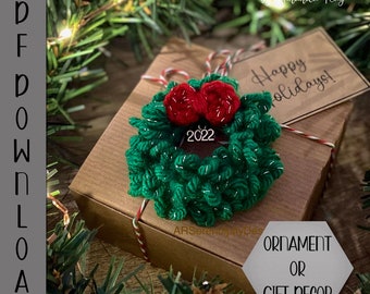 May include: A green yarn wreath with a red bow and silver glitter, with the year 2022 in the center. The wreath is on a brown box with a white tag that says "Happy Holidays!" and a white hexagonal tag that says "ORNAMENT OR GIFT DECOR".