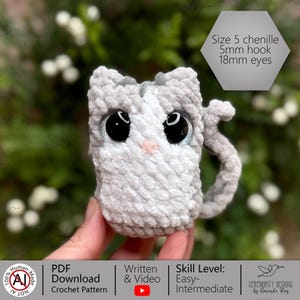 K&ouml;nnte beinhalten: Eine handgefertigte, graue, geh&auml;kelte katzenf&ouml;rmige Tasse mit gro&szlig;en schwarzen Augen und einer rosa Nase. Die Tasse hat einen Henkel und ist aus Chenille-Garn der Gr&ouml;&szlig;e 5 gefertigt. Der Text auf dem Bild lautet "Size 5 chenille, 5mm hook, 18mm eyes."