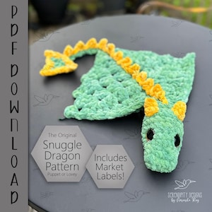 May include: A handmade green crochet dragon puppet or lovey with yellow accents. The dragon has a textured body, yellow spines, and small black eyes. The image includes text that reads "The Original Snuggle Dragon Pattern" and "Includes Market Labels!"