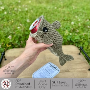 May include: A crocheted whale-shaped can cozy in shades of gray, holding a red soda can. The cozy has a black eye and a white belly. The image also shows a tag with text and the words "PDF Download Crochet Pattern".