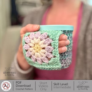 May include: A crocheted mug cozy with a floral design in pastel colors, held in a hand. The cozy features a square granny stitch flower in shades of lavender, cream, and yellow, framed by a mint green border. The mug is a light teal color. The image includes text that says "PDF Download Crochet Pattern".