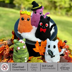 Może przedstawiać: Kolekcja ręcznie robionych szydełkowych figurek na Halloween. Figurki obejmują czarnego kota z czerwoną muszką, fioletową wiedźmę, pomarańczowego kota, zielonego zombie i białego ducha trzymającego rybę. Figurki są wystawione na czarnym kotle.