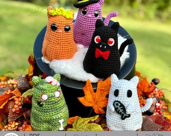 Puede incluir: Una colección de figuras de Halloween hechas a mano con ganchillo. Las figuras incluyen un gato negro con pajarita roja, una bruja morada, un gato naranja, un zombi verde y un fantasma blanco con un pez. Las figuras se exhiben en un caldero negro.