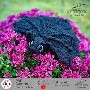 Può includere: Un pipistrello all'uncinetto nero con grandi occhi neri lucidi, ali testurizzate e orecchie arrotondate, appoggiato su un denso grappolo di fiori di crisantemo color magenta brillante. L'immagine include il testo Serendipity Designs by Amanda Ray.